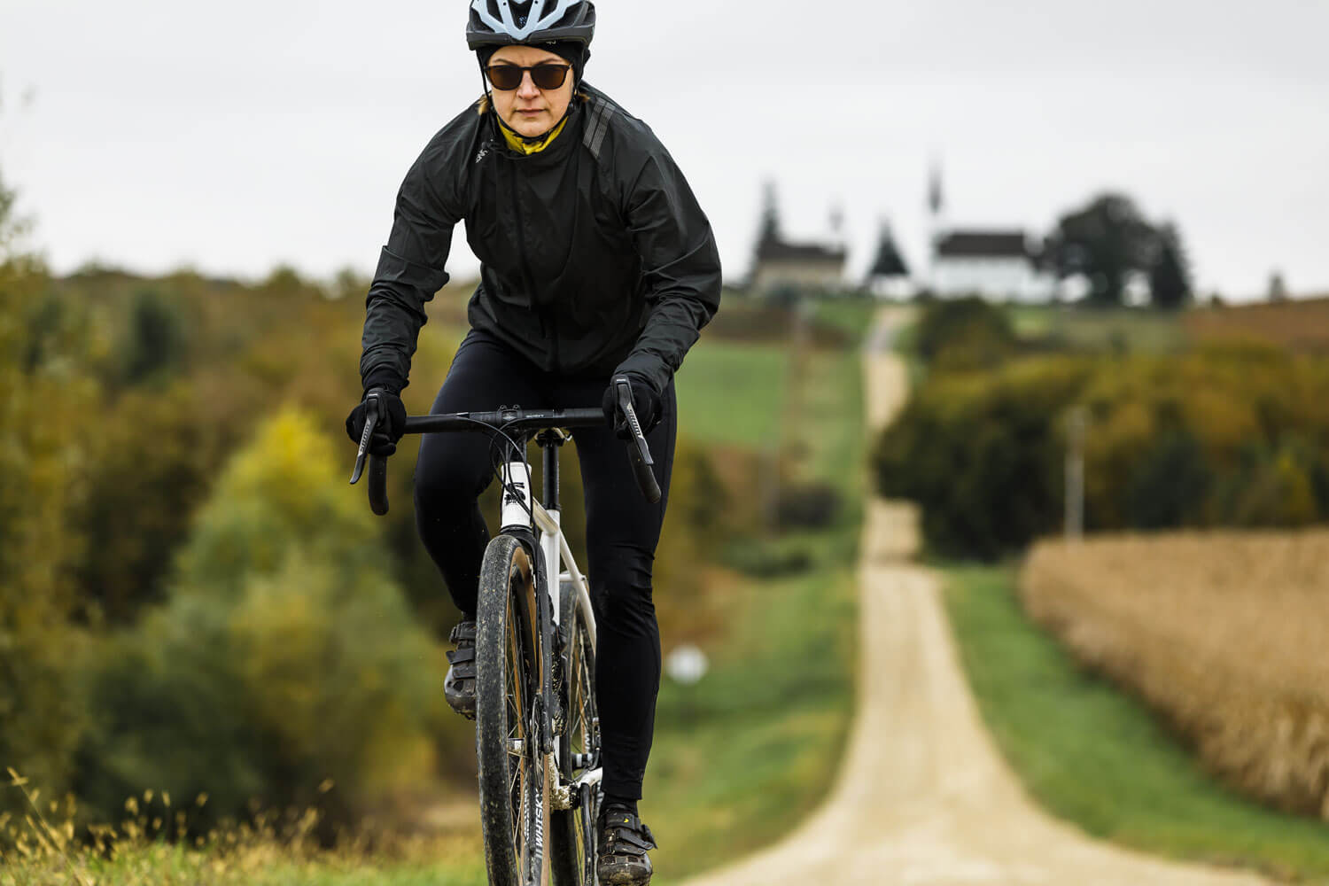 Shown from the front, a cyclist wearing all black and gloves rides on down a gravel path.