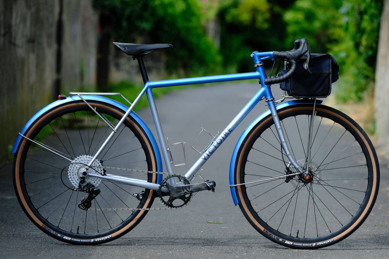 Profile view of Victoire Cycles blue randonneur bike with a handlebar bag. A blurry paved road is in the background.