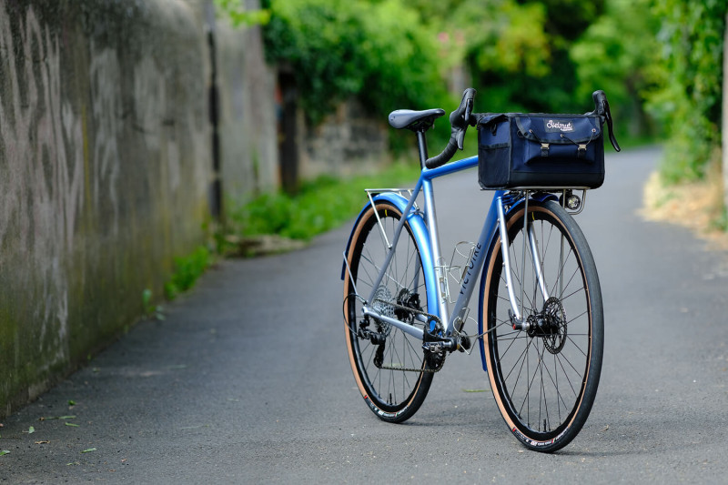 Front three quarter view of Victoire Cycles blue randonneur bike with a handlebar bag on a paved road.