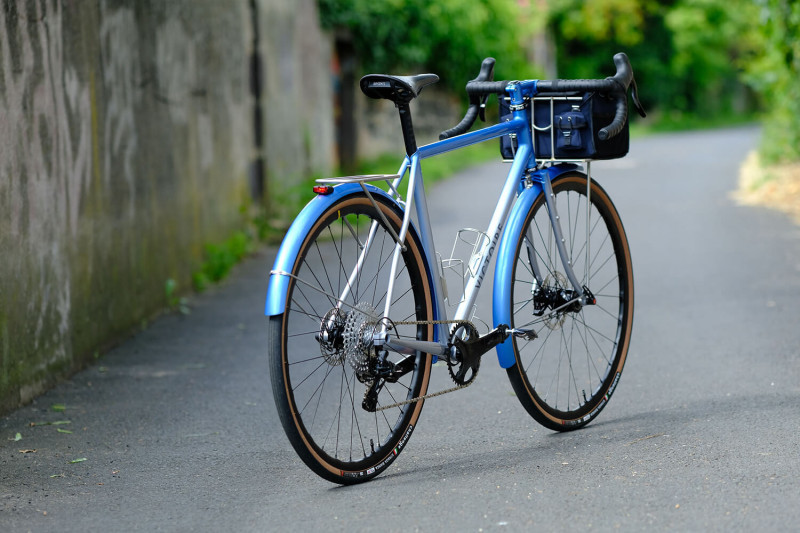 Rear three quarter view of Victoire Cycles blue randonneur bike with a handlebar bag on a paved road.